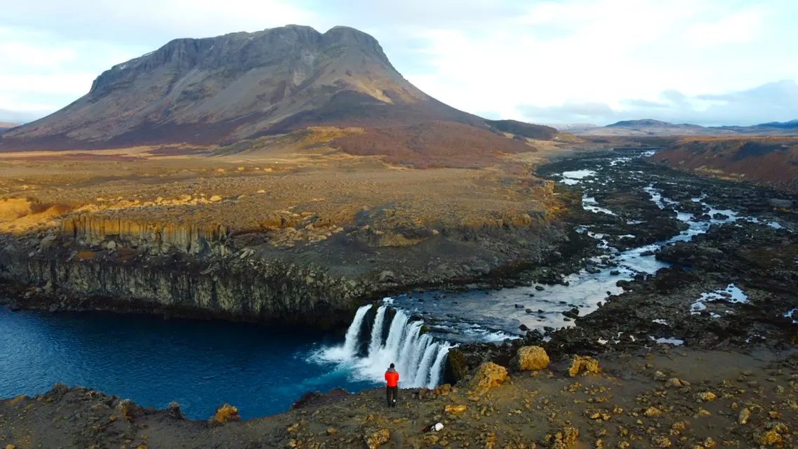 A photographer beside a waterfall beneath a mountain in Iceland